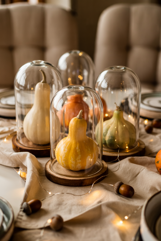 Elegant Thanksgiving centerpiece of glass cloches highlighting cream, orange, and green gourds on a linen runner, with fairy lights, acorns, ivory plates, and crystal glasses creating a cozy, curated fall tablescape.