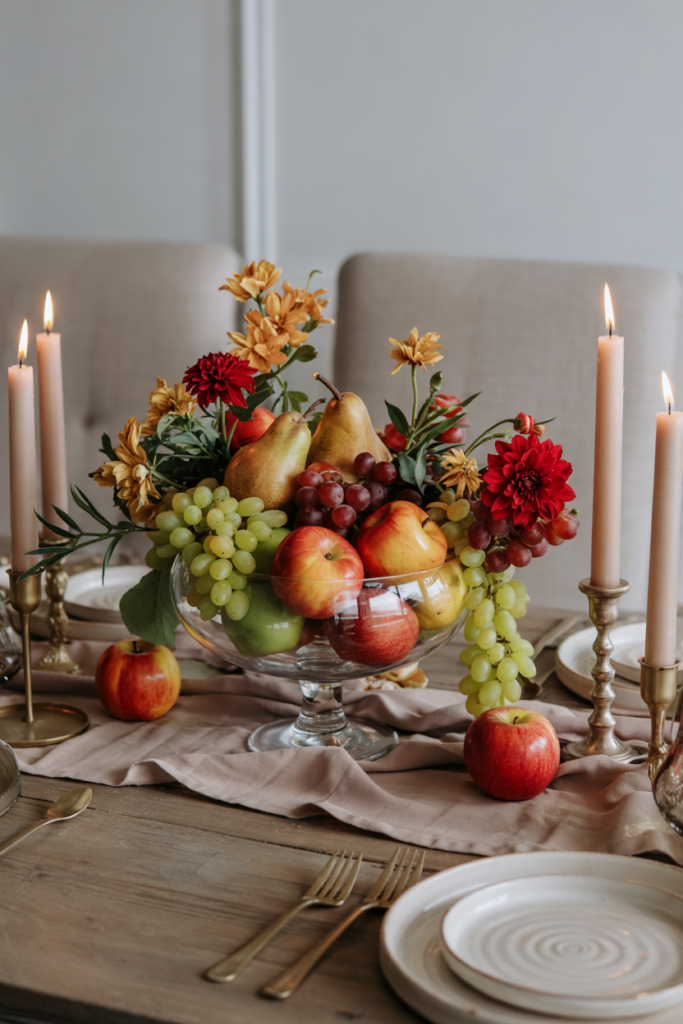 Abundant Thanksgiving table centerpiece with apples, pears, grapes, dahlias, and chrysanthemums in a compote bowl, styled with gold candlesticks, pale ceramic plates, gray chairs, bread rolls, and honey butter for warmth.