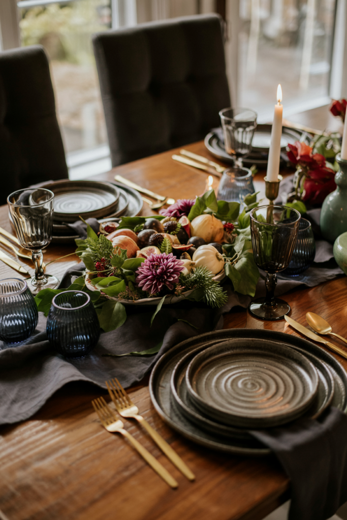 Elegant jewel-tone Thanksgiving table featuring emerald greenery, plum dahlias, sapphire glass votives, black chargers with ivory plates, smoked-glass goblets, gold flatware, and a platter of roasted root vegetables and figs.