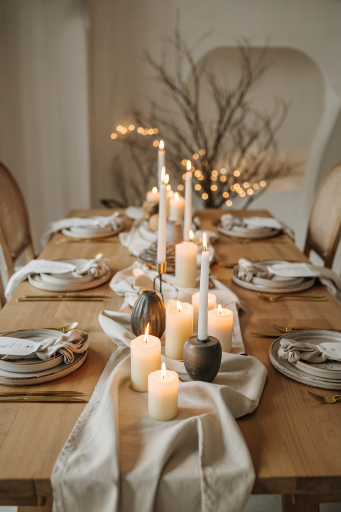 A minimalist monochrome neutral linen runner in layered oat, camel and bone tones stretches down a pale oak table and is topped with clusters of pillar and tapered candles in varied holders, textured linen napkins at each setting and clear glassware