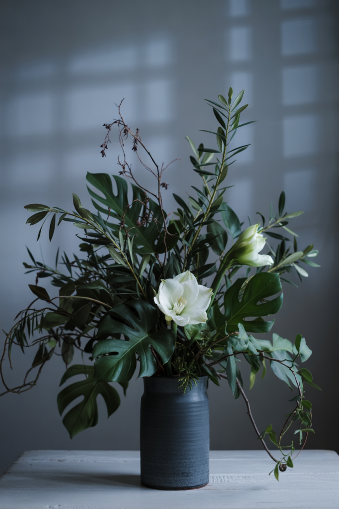 Sculptural foliage-focused display of large leaves, monstera ,cascading ivy and two ivory accent blooms in a tall matte vase.