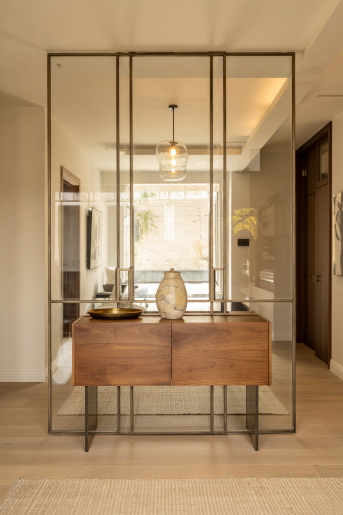 Luxury glass and metal entryway with framed glass partition, warm wood console, and airy modern design.