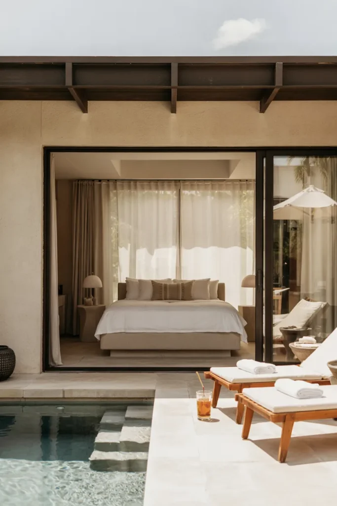 Monochrome minimalist pool house guest suite in beige and black tones with open glass doors leading to a calm summer pool, styled like a luxury resort retreat.